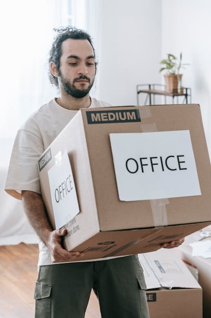 A person is seen packing a cardboard box with a black electronic device, possibly a computer or gaming console, inside a room with a wooden floor. The individual is wearing a light-colored short-sleev
