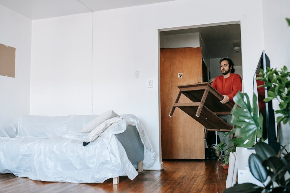 A professional mover from Storage Merton is carrying a vintage-style green velvet sofa with wooden legs during the home relocation process inside a room with white walls, a wooden ceiling with exposed