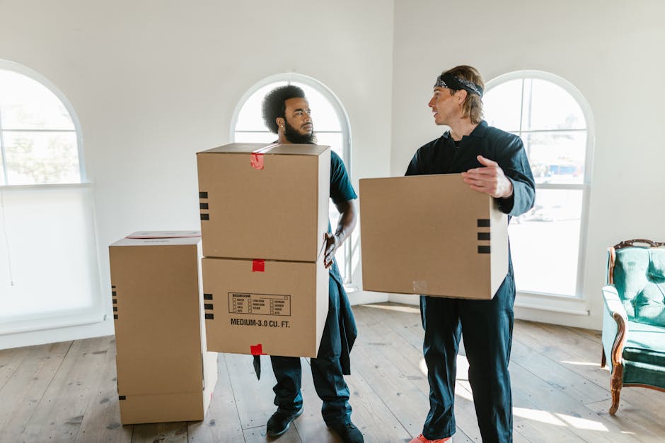Two professional movers from Storage Merton are sitting inside the open rear of a white moving van, which is parked on a paved street under a cloudy sky. The interior of the van contains some packing